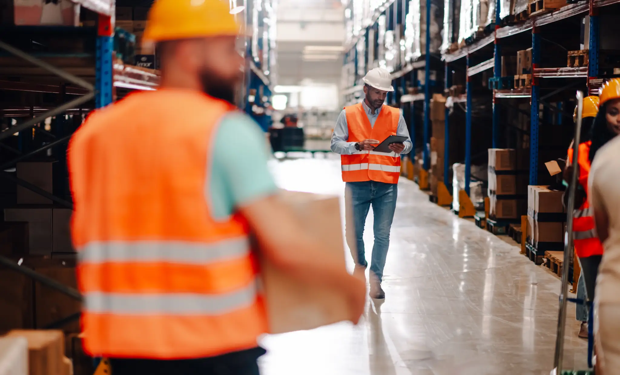 Warehouse manager walking through the aisle, using a digital tablet to monitor operations, while other workers carry boxes in a spacious, modern distribution center
